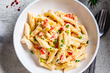 Penne pasta with chicken, pepper and green onions in creamy sauce in a white plate, gray background, top view.