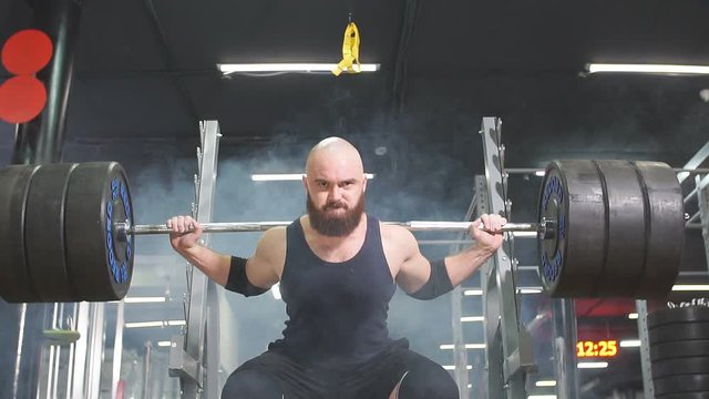Young strong bodybuilder preparing for competition of powerlifters, doing squats using heavy metal barbell training hardly in underground garage