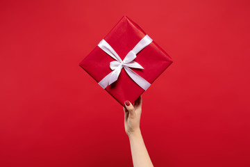 partial view of woman holding wrapped Christmas present with white ribbon in hand isolated on red background. Christmas concept