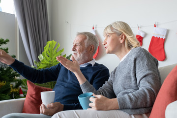 senior elderly caucasian old man and woman drinking coffee with reading book and sitting on sofa and discussion together in living room that decorated for happy christmas festival day in the morning