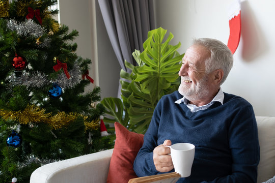 senior elderly caucasian old man drinking coffee with reading book and sitting on sofa in living room that decorated for happy christmas festival day in the morning