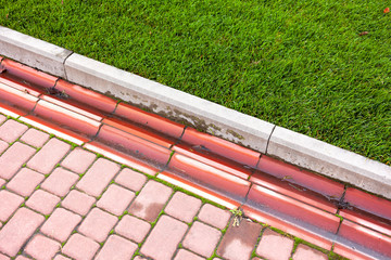 Drainage system in the park by the green lawn and footpath of red paving slabs. - obrazy, fototapety, plakaty