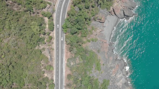 A flying camera takes a motorcyclist passing on a road near the sea with a view of the rocky shore.