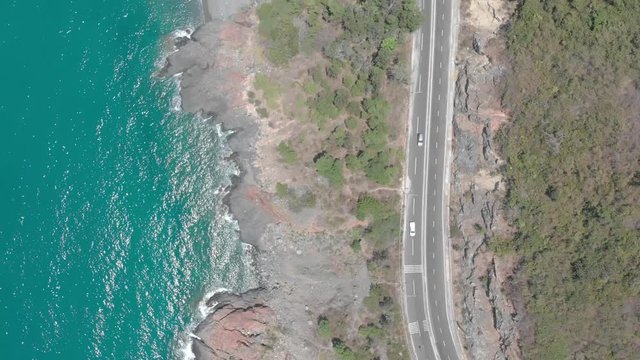 A flying camera takes a motorcyclist passing on a road near the sea with a view of the rocky shore.