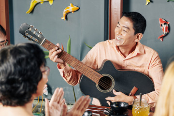 Happy middle-aged Asian man playing guitar and singing a song for his relatives at family dinner