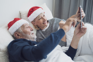 happy elderly couple caucasian senior man and woman with red hat talking family by using on tablet for christmas festival day in the morning in bedroom, retirement love family lifestyle concept