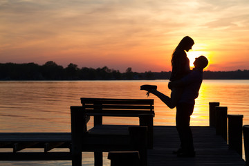 Fototapeta na wymiar Romantic male and female couple lift and embrace on a dock, backlit by a sunset