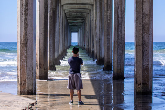 A young man taking pictures of the ocean from under concrete pier, San Diego, California