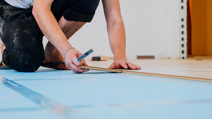 A worker is fitting laminate with a piece of a panel and a small hammer in the room. A person is sitting on a knee and moving along the laminate line, side view, legs and arms in shot.