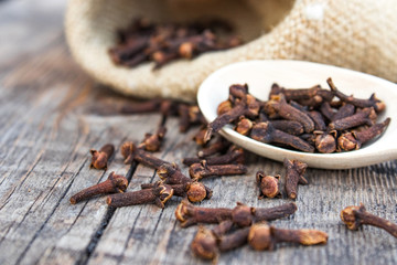 A spice of dried cloves lies on a wooden spoon and is scattered on old wooden boards near the bag.