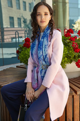 Medium full shot of a brunette lady with a black handbag in a pink coat sitting on a bench in urban...