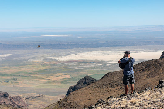 A man standing on a side of Steens Mountains Summit taking pictures of underlying desert