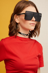 Close-up shot of a young chestnut-haired European girl in a red tee, golden earrings, black choker with crystal lettering and square oversized sunglasses with black rim and black lenses.