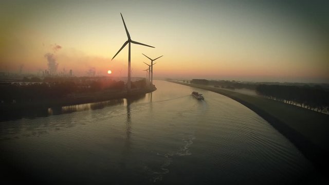 Drone shot of wind turbines at sunset with a river and a ship. Petrochemical industry visible in background. Pernis.