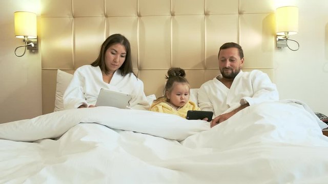 Mom watches social networks in a tablet, dad shows little daughter cartoons on a smartphone in bed