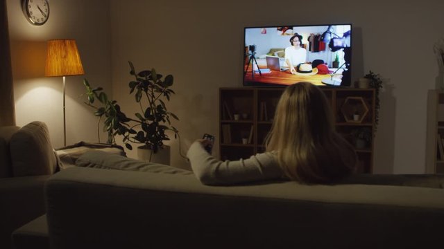 Tracking with rear view of unrecognizable woman sitting on couch in living room lit by lamplight and changing channels on TV
