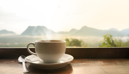 White ceramic coffee cup on wooden table  in morning with sunlight over blurred mountains landscape 