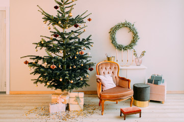 Decorated cristmas tree with gift boxes, chair, puff and table with wreath hanging on wall. Winter holidays interior.
