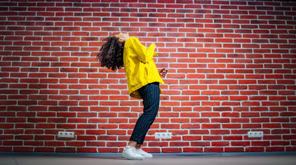 Positive human emotions. Portrait of happy emotional teenage girl with curly hair bending back, view from the side. Wearing bright yellow cotton jacket and white shirt. Brick wall background.