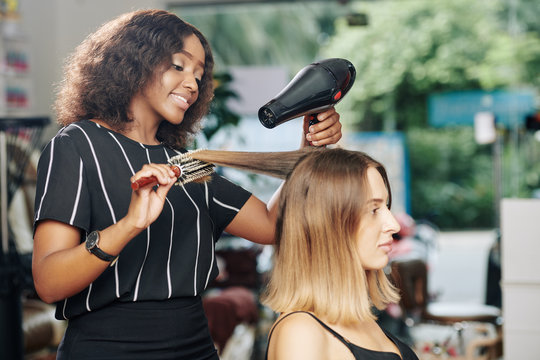 Positive young Black hairdresser enjoying working in beauty salon and blowdrying hair of client