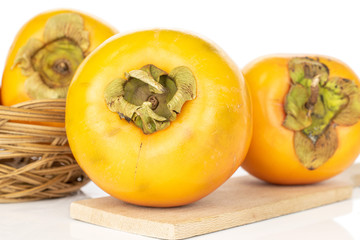 Group of three whole sweet orange persimmon in round rattan bowl on wooden cutting board isolated on white background