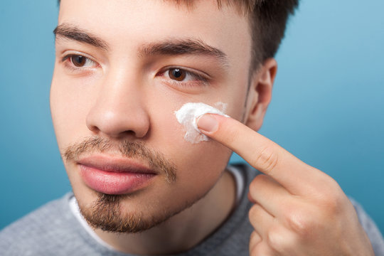 Male beauty and skincare. Portrait of young handsome man with small beard and mustache applying cream or anti-aging cosmetics on face, looking aside. indoor studio shot isolated on blue background