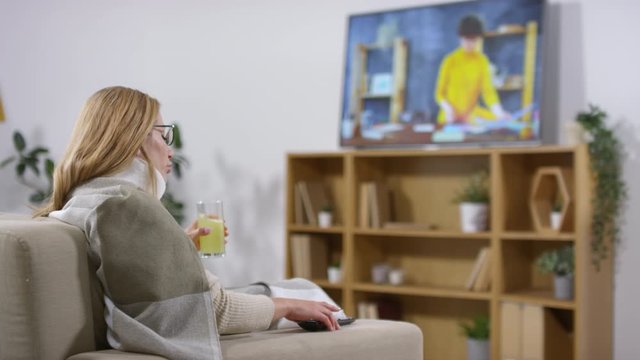 Medium shot of sick woman sitting on couch and drinking hot remedy while changing TV channels