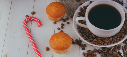 A hot cup of coffee with the coffee beans, small cupcakes and a candy on the table. Composition of a delicious coffee and sweet snacks.
