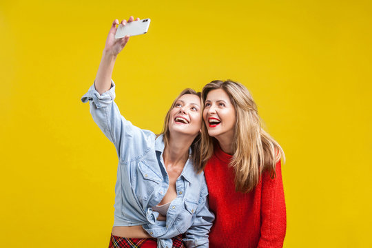 Female friends enjoying time. Portrait of two beautiful women in stylish clothes standing with toothy smile and taking selfie on phone, happy memories. indoor studio shot isolated on yellow background