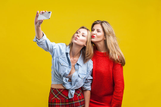 Female friends sending kiss while taking selfie. Portrait of two attractive women in stylish clothes standing, using smartphone to take funny photo. indoor studio shot isolated on yellow background