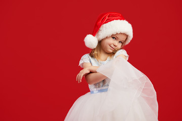 A little girl in a white princess dress and Santa hat holds snowflakes in her hands and looks at them. Red isolated background. The concept of New Year and Christmas holidays
