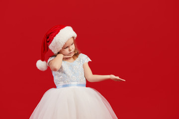 Little girl in a white princess dress and Santa hat shows with his hand to the right on an empty place for text. Red isolated background. The concept of New Year and Christmas holidays