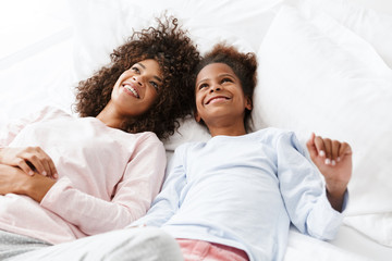 Image of cheerful african american woman and her daughter lying in bed