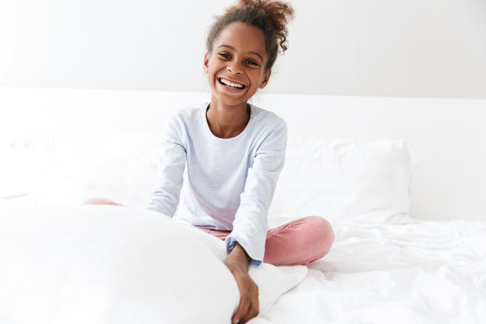 Image of african american little girl smiling and sitting in bed at home