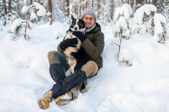 Happy man holding lovely dog in his hands in snowy forest. Smiling boy hugging adorable puppy in winter wood. Pet lover.  Dog - human`s friend concept.