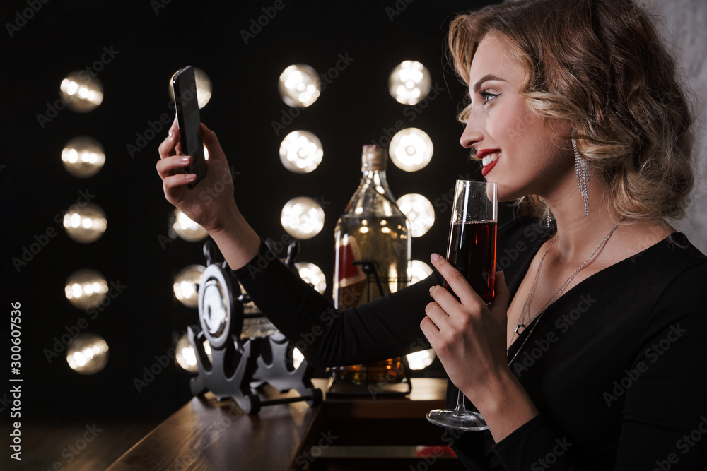 Poster image of young blonde woman using cellphone and drinking red wine in bar