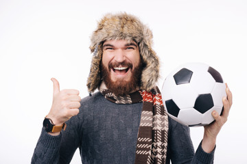 Cheerful bearded hipster man holding soccer ball and showing thumbs up