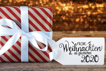Fototapeta na wymiar One Christmas Gift With Label With German Calligraphy Frohe Weihnachten Und Ein Glueckliches 2020 Means Merry Christmas And A Happy 2020. Wrapping Paper With A Bow And Lights In Background