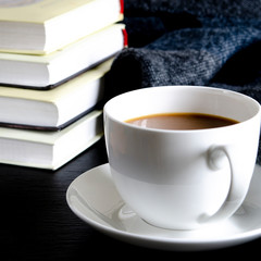 A white cup with a fragrant cove stands on a black table next to books and a blue wool scarf