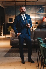 Mature handsome businessman stand and hold glass of whiskey in his own office.