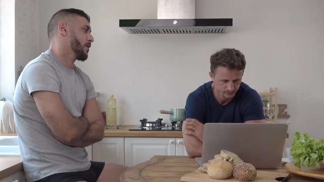Young fit masculine handsome gay male couple acting concerned impatient and frustrated while using on notebook computer at kitchen table