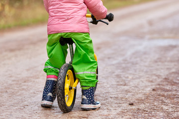 Rear view of low section of a child girl sitting on her balance bicycle in warm and waterproof clothing on a wet and dirty gravel road in autumn. Seen in Franconia / Bavaria in Germany in November.