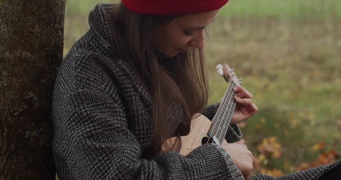 Close up portrait of hipster girl sitting with ukulele on ground outdoors on nature background. Young woman in red hat learning how to play Hawaiian guitar slow motion. Musical tutorial education