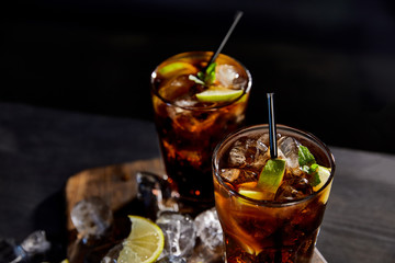 cocktails cuba libre in glasses with straws, ice cubes and limes on black background