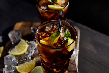 selective focus of cocktails cuba libre in glasses with straws, ice cubes and limes on black background