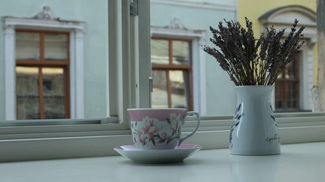 A cup of hot tea and a vase with dry lavender flowers are on the windowsill against the background of the house opposite