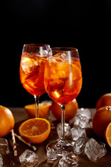 Aperol Spritz in glasses, oranges and ice cubes on black background