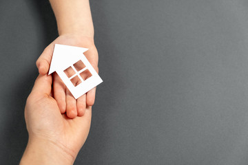 Close up horizontal image of female and child hands holding white paper house on gray background. Family home and real estate concept. Flat lay, copy space