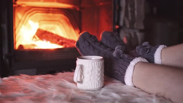 Feet in woollen socks by Burning Christmas Fireplace. 4K STABILIZED shot. Woman relaxes by warm fire with a cup of hot drink and warming up her feet. Close up. Winter and Christmas holidays concept.