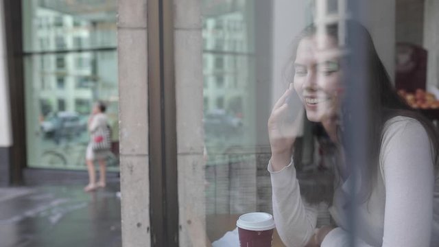 Young woman in a cafe enjoying a coffee and using a smartphone - Beautiful brunette sitting behind a window in a cafe bar in London, looking away from camera - Lifestyle and food drink concepts